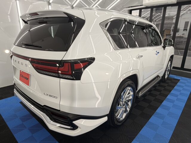 Rear three-quarter view of a white Lexus LX600 SUV parked indoors on a blue-and-black tiled floor in a showroom.