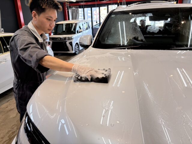 Man in a gray uniform cleaning a white car hood with a soapy sponge inside a showroom.