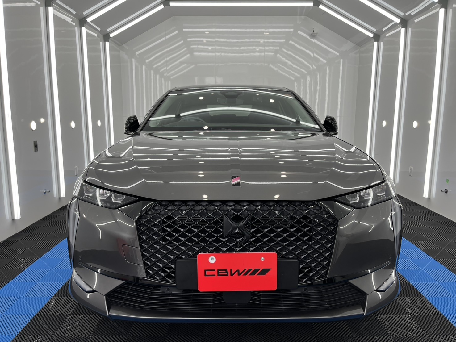 Front view of a gray SUV with a bold diamond-pattern grille in a bright white showroom with vertical LED accents on the walls.