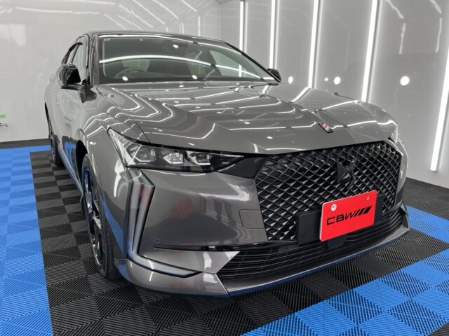 Front three-quarter view of a gray SUV with a bold diamond grille in a studio, blue-and-black checker floor, red license plate area reading CBWW.