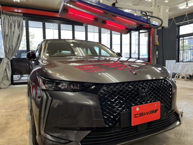 Grey luxury SUV/ sedan inside a showroom with a large black mesh front grille and red license plate, under overhead red thermal lamps.