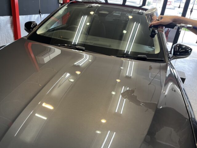 Gray car hood in a workshop as a technician uses a spray gun for detailing near the windshield.
