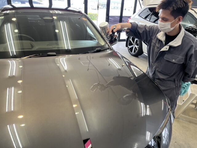 Auto detailer wearing a mask polishes the hood of a gray car with a polishing machine in a showroom.