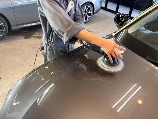 Person in mechanic coveralls polishes a car hood with a polishing machine, creating a shiny, even finish in a workshop.
