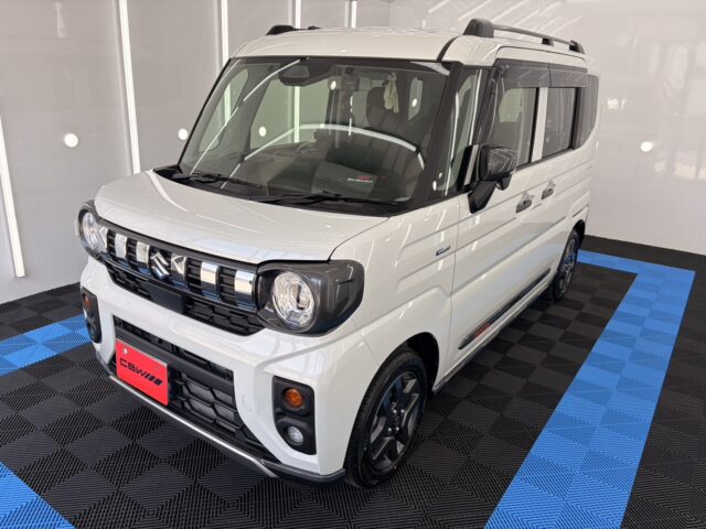 Front three-quarter view of a white Suzuki minivan in a showroom, glossy paint, black grille and round headlights, blue-and-black patterned floor beneath the car.