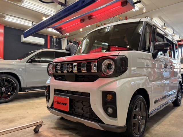 White compact van in a showroom foreground, with a black grille and red license plate, and a silver SUV beside it in a workshop setting.