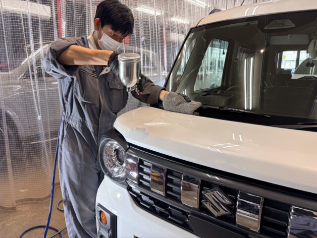 Auto detailer in mask and gray coveralls polishing the white van's hood with a spray gun in a workshop environment.