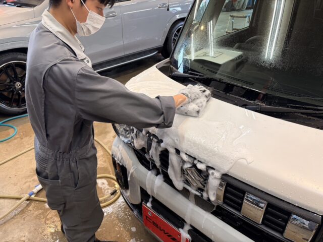 Detailer wearing a mask scrubs the hood of a white car with soapy foam in a garage setting.