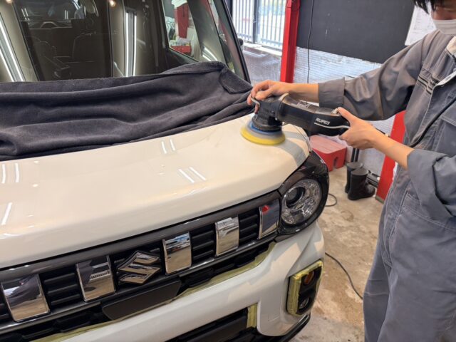 Professional detailer polishes the white car hood with a handheld buffer in a workshop setting, using a yellow pad on the surface.