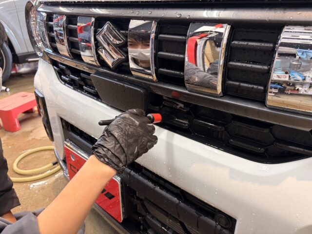 Technician with black gloves using a red-handled tool on the front bumper/grille of a Suzuki SUV in a workshop.
