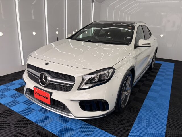 Front three-quarter view of a white Mercedes-Benz SUV (AMG) in a white photo studio on a blue-and-black checkered floor, showing the grille and emblem clearly.