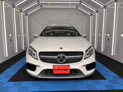 Front view of a white Mercedes‑Benz SUV facing the camera, inside a bright white studio with vertical LED accents and a blue-and-black checkered floor.
