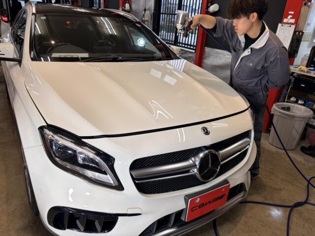 Auto technician in gray coveralls polishes the hood of a white Mercedes SUV with a spraying tool in a workshop.