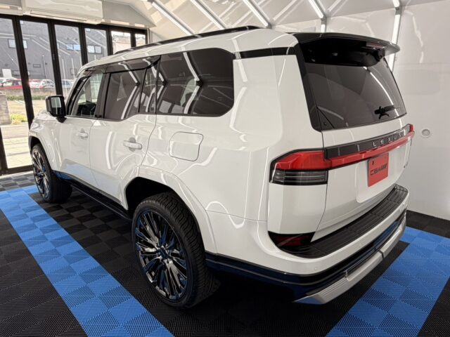White Lexus SUV in a showroom, rear three-quarter view, tinted windows and large chrome wheels on a blue/black checkered floor.