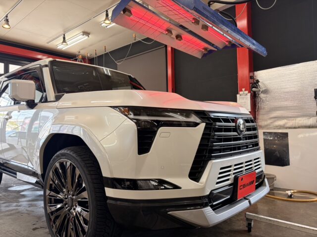 Front three-quarter view of a white luxury SUV inside an auto service bay, with a red overhead heater and red support columns visible above the vehicle's hood.