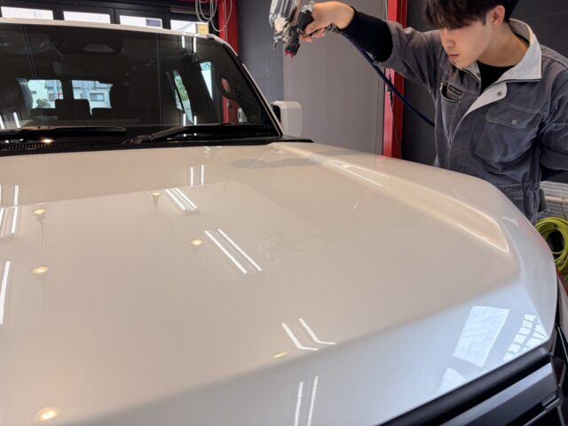 Auto body technician in gray coveralls spraying a white car hood in a workshop with a paint gun overhead period.