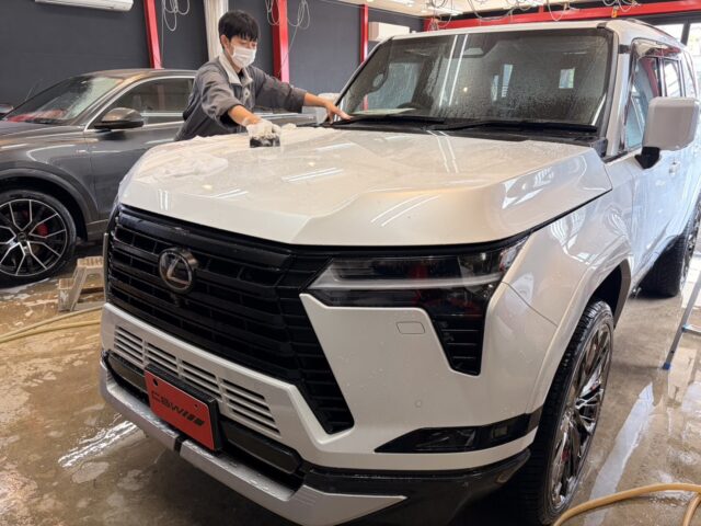 Worker wearing a mask washes the white SUV inside a car detailing shop, wiping the hood with a cloth.