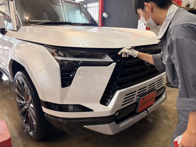 Technician wearing a mask and gloves detailing the front grille of a white SUV in a workshop.