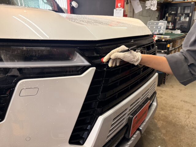 Automotive technician in gloves using a tool on the front grille of a white vehicle in a repair shop.