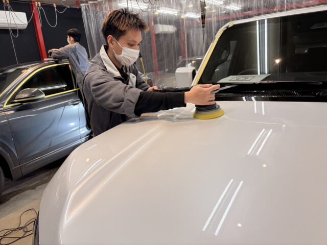 A worker wearing a mask polishes the hood of a white SUV with a power buffer in an auto shop, another car in the background.