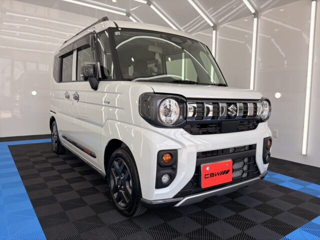 White Suzuki compact van in a bright showroom, facing the camera with round headlights and a bold grille.
