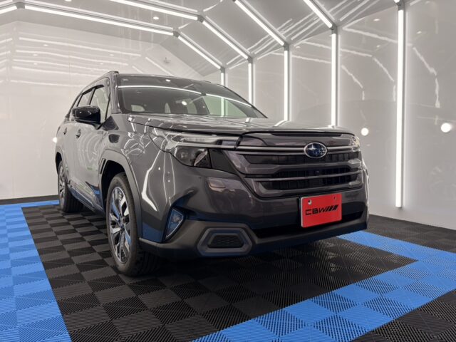 Gray Subaru SUV in a bright showroom with white walls and neon lighting, front three-quarter view showing grille and wheel rims with a red license plate area.