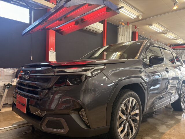 Front three-quarter view of a dark SUV inside a workshop, with a red overhead heater and workshop equipment nearby.
