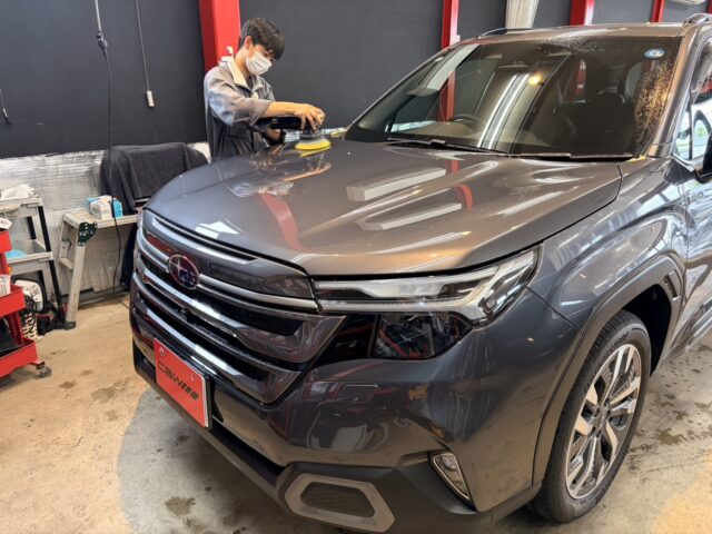 Auto detailer wearing a mask polishes the hood of a gray SUV with a handheld polisher inside a workshop.