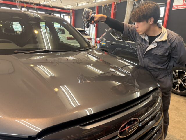 Detailer in gray coveralls sprays the hood of a gray Subaru SUV with a detailing gun in a professional auto shop.