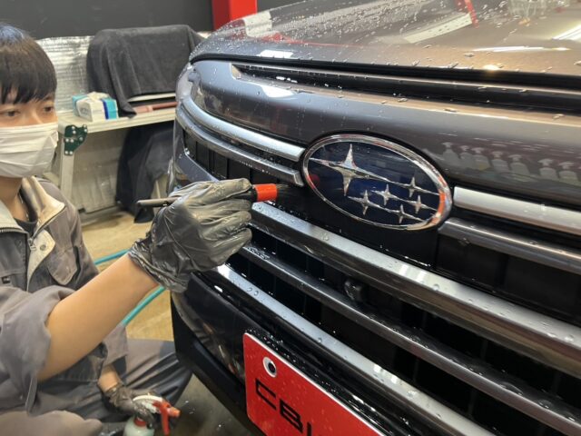 Auto technician wearing a mask and gloves applies a tool to the car's front grille, likely detailing or applying coating in a workshop.