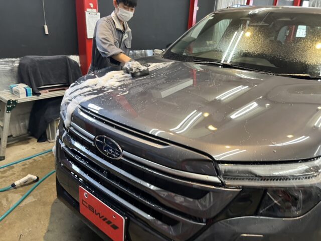 Worker wearing a mask and gloves washes a gray SUV in a garage, suds on the hood and windshield visible.
