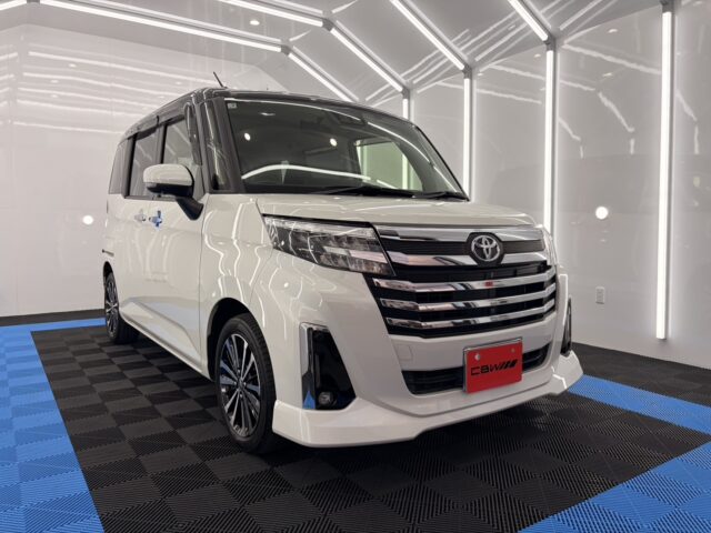 White Toyota minivan in a bright showroom with a black grille and chrome accents; front angled view shows blue floor mats beneath.