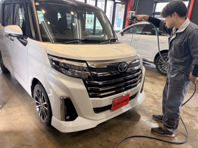 Technician in gray coveralls using a spray gun to wash a white Toyota minivan in a car service bay.