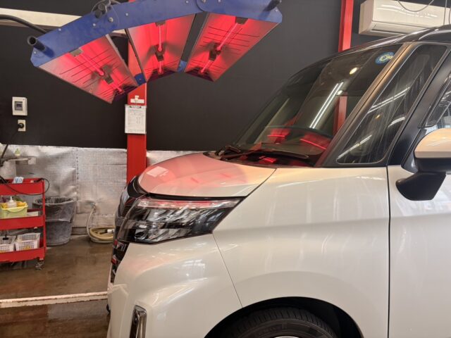White van inside a car service bay under red infrared lamps, with a tool cart and workshop equipment nearby.