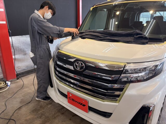 Auto technician wearing a mask polishes the hood of a white Toyota van in a workshop, with masking tape and a towel on the windshield.