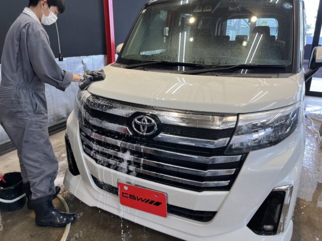 Worker in gray coveralls and mask washing the front of a white Toyota van with soapy suds in a car wash bay