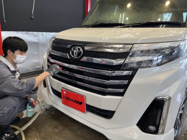 Worker wearing a mask, washing the front grille of a white Toyota van with a hose in a service bay.