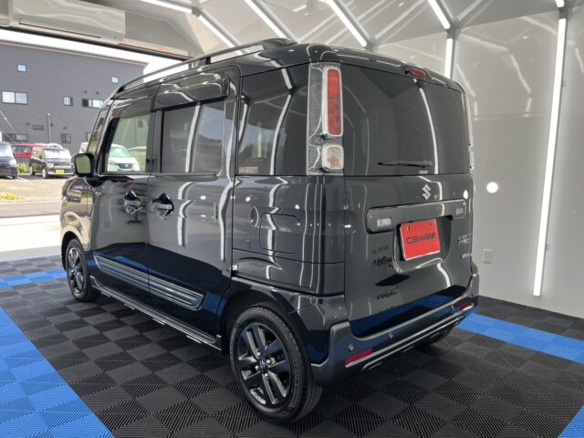Rear three-quarter view of a dark gray Suzuki compact van in a showroom, displaying glossy panels and blue-and-black checkered flooring.