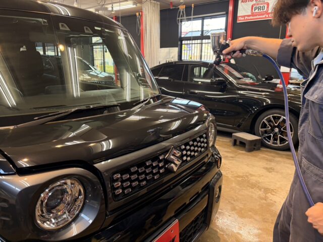 Technician using a spray gun to apply coating on the front of a black SUV in an auto detailing shop, with another black car in the background.