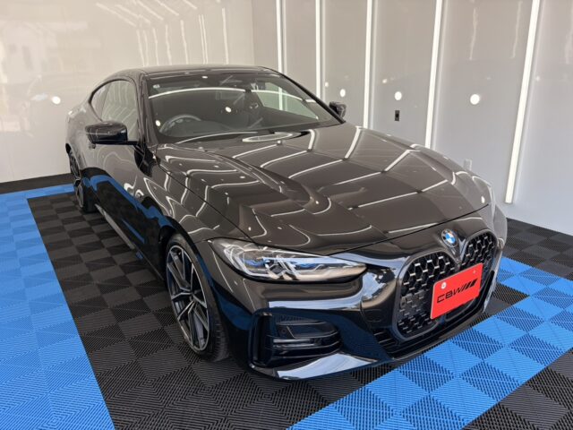 Black BMW sedan on a blue-and-black checkered showroom floor, front three-quarter view with glossy finish and reflections.