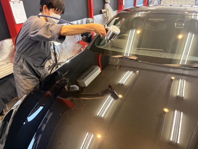 Auto detailer wearing a mask and gray coveralls polishes the hood of a dark car in a shop stall.