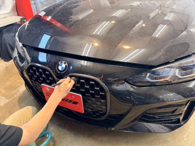 Person detailing a black BMW, applying a treatment to the front grille with a tool; water droplets on the hood and reflections from overhead lights nearby.