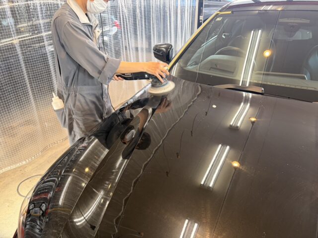 Technician in coveralls polishing a car hood with a handheld polishing machine, wearing a mask in a workshop.