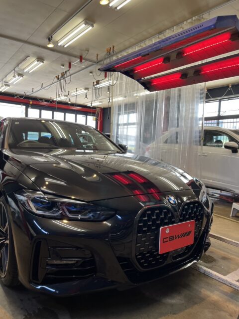 Black BMW SUV in an auto service bay, front grille and glossy hood visible under bright shop lights and red overhead heaters, red plate on the front.