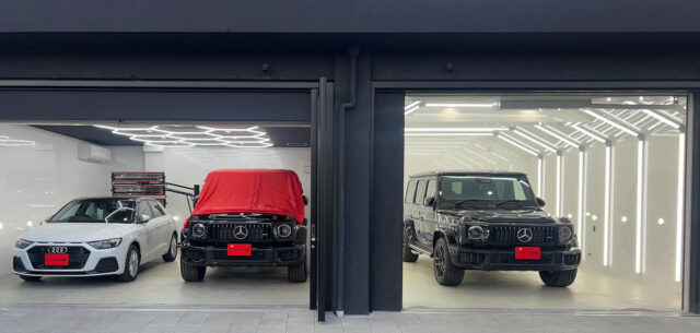 Showroom with two lit garage bays: left white Audi sedan with a roof rack, center black Mercedes G‑Wagon partially covered in red cloth, right black Mercedes G‑Wagon in a bright white space