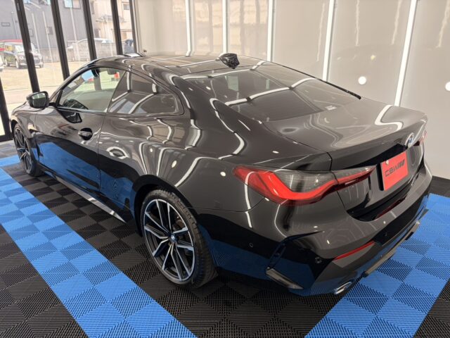Rear three-quarter view of a dark gray BMW coupe in a showroom, highlighting its sleek lines and red taillights on a blue-and-black tiled floor.