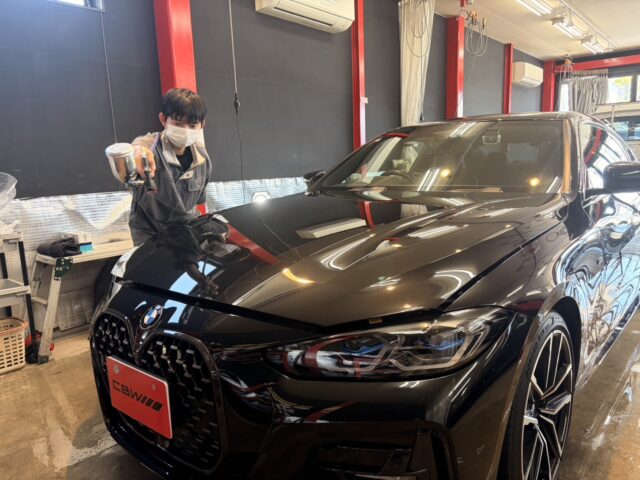 Detailer in a mask applying spray coating to the hood of a glossy black BMW in a workshop