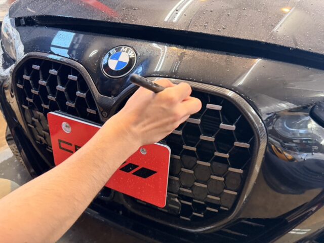 Person cleaning the front grille of a dark BMW with a detailing tool in hand, water droplets on the hood nearby.