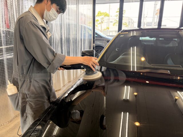 Auto detailer wearing a mask polishing a black car hood with a handheld polisher in a bright workshop is visible in the image.
