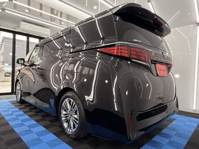 Rear three-quarter view of a dark gray Toyota Alphard minivan inside a showroom, reflecting neon lights on glossy body.